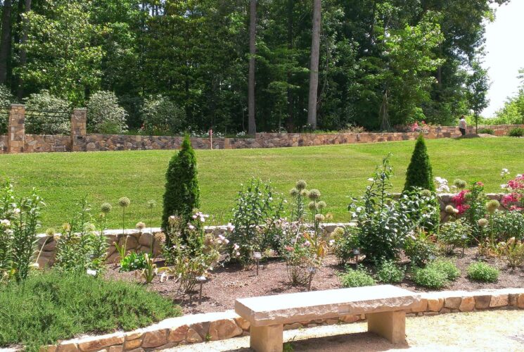 Stone brick flower garden full of colorful flowers next to large green lawn with large green trees in the background