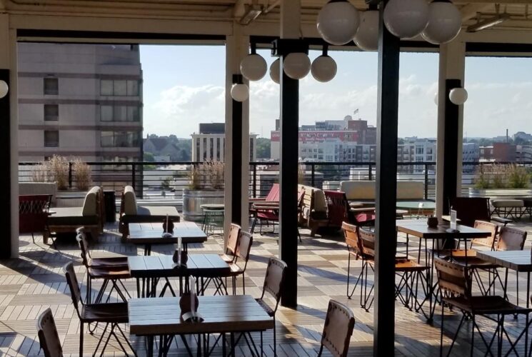 Large room in a restaurant with wooden and wrought iron tables and chairs and an open air side with a downtown view