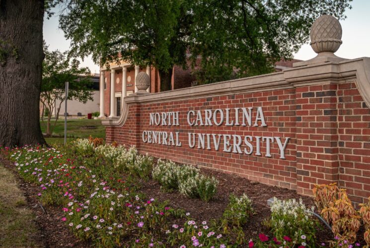 Red brick sign with North Carolina Central University letters in white next to a colorful flower garden and large tree