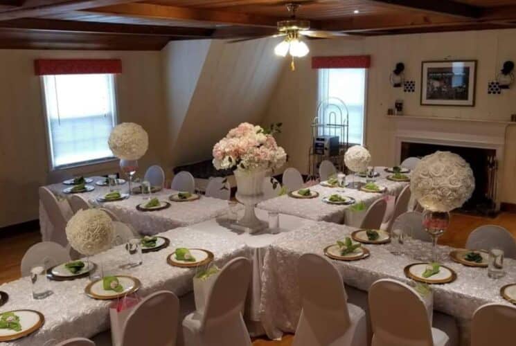 Large room with white panel walls, hardwood flooring, fireplace, and multiple rectangle tables and chairs covered with white tablecloths and covers