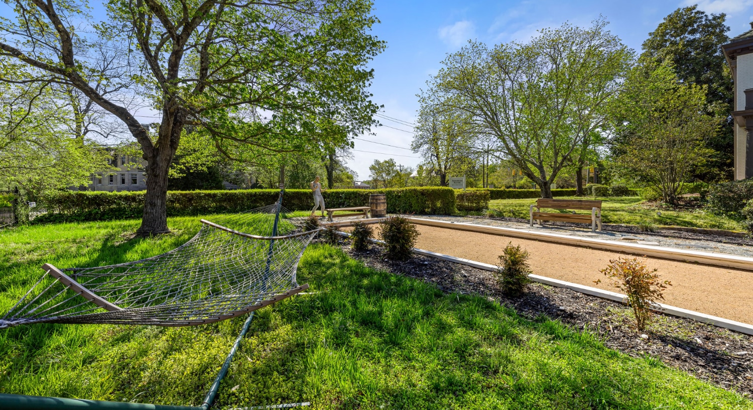 Large backyard with green grass, hammock, bocce court, green shrubs, and large trees