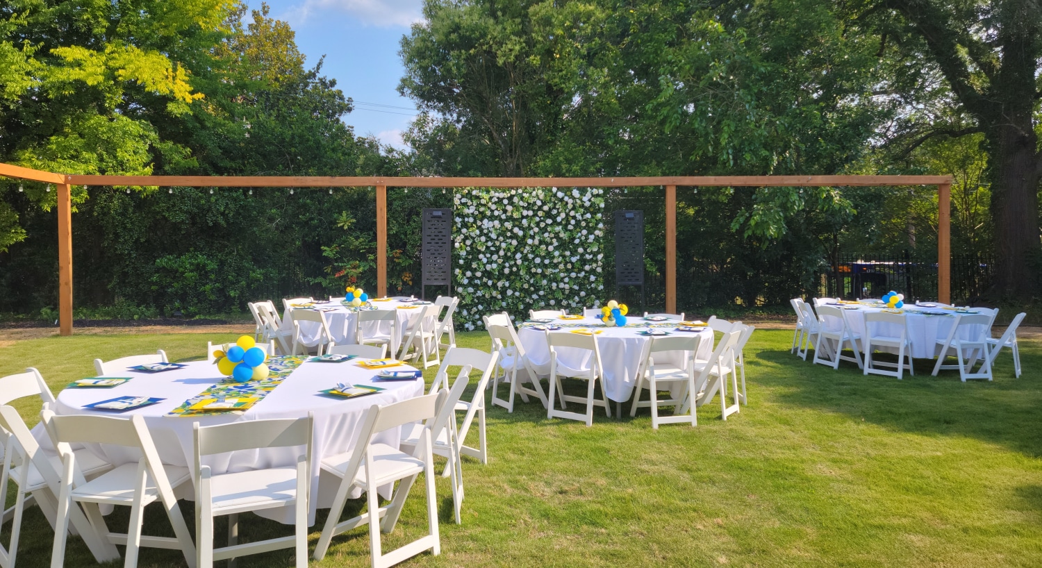 Backyard space set up for an event with multiple round tables with white tablecloths and white chairs set up on a green lawn surrounded by large trees