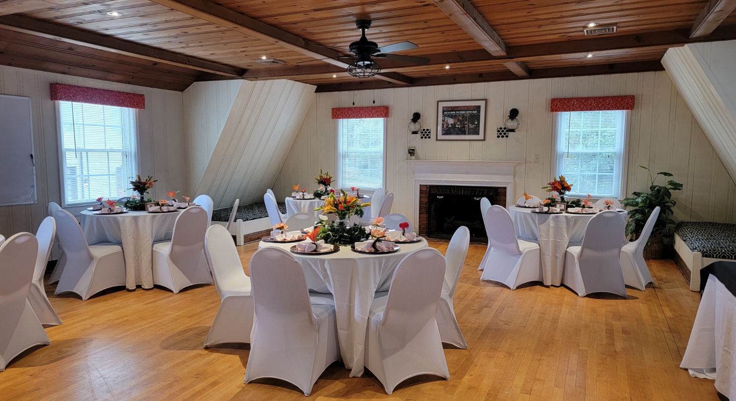 Large room with white panel walls, hardwood flooring, fireplace, and multiple round tables and chairs covered with white tablecloths and covers