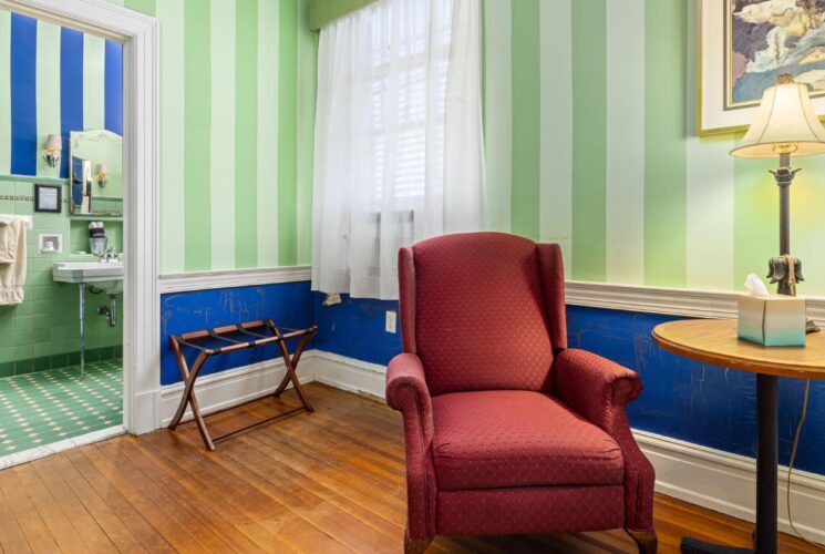 Bedroom sitting area with light green striped walls, blue wainscotting with white trim, red upholstered armchair, small round table, and view into bathroom