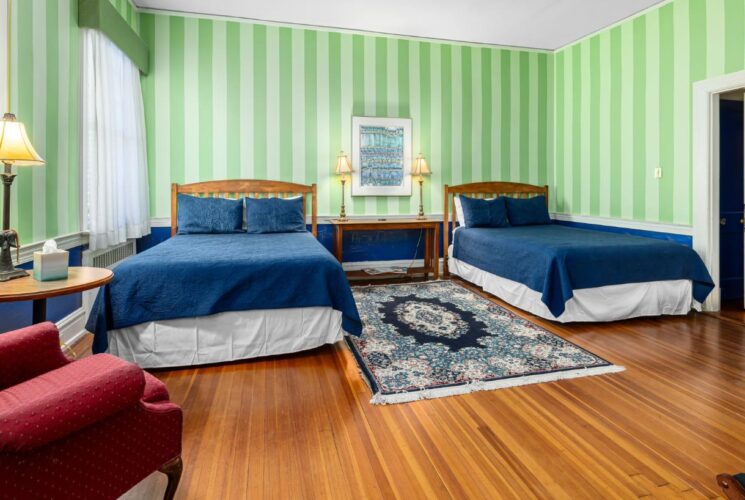 Bedroom with light green striped walls, blue wainscotting with white trim, hardwood flooring, wooden bed, navy blue bedding, red upholstered armchair, and small round table