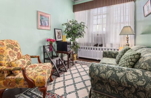 Room with light green walls, hardwood flooring, green floral upholstered sofa, yellow floral upholstered antique chair, and large windows