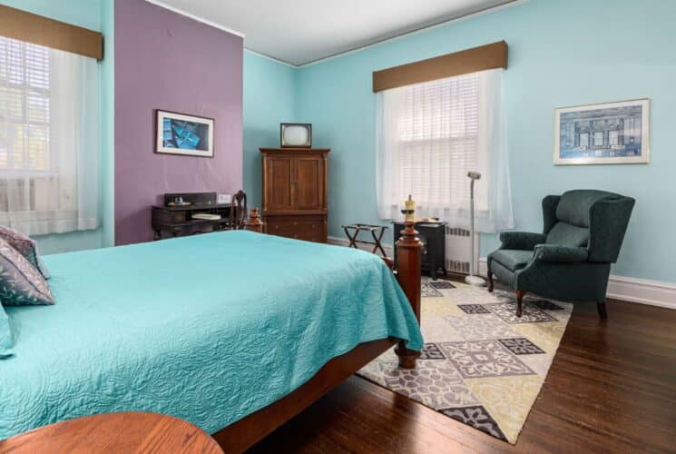 Bedroom with light blue and purple walls, hardwood flooring, wooden bed, turquoise bedding, dark wooden antique desk, and green upholstered armchair