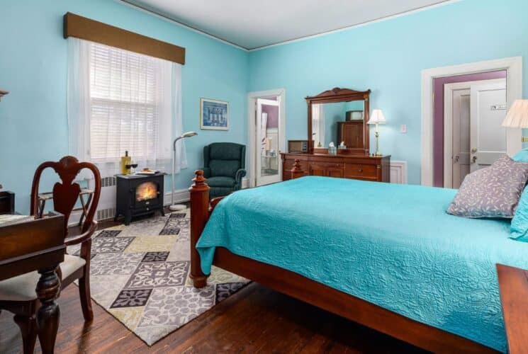 Bedroom with light blue and purple walls, hardwood flooring, wooden bed, turquoise bedding, dark wooden antique desk, and green upholstered armchair