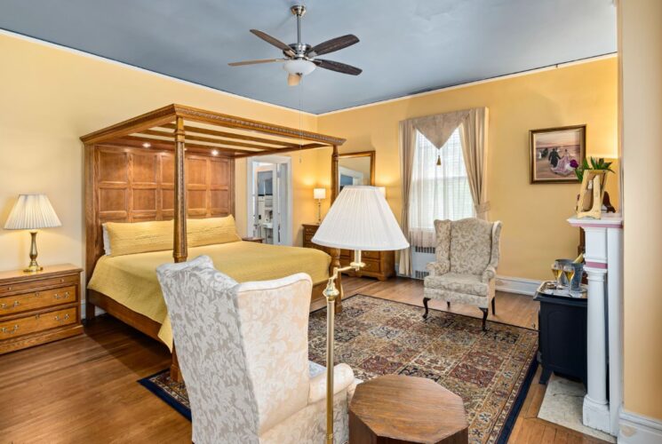 Bedroom with apricot walls, hardwood flooring, wooden four-poster canopy bed, yellow bedding, wooden dresser, and sitting area