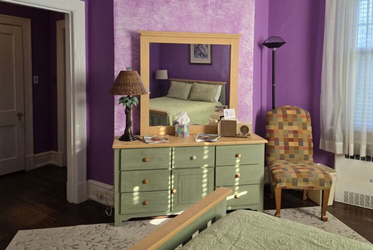 Bedroom with purple walls, hardwood flooring, wooden bed, light green bedding, and light green wooden dresser with mirror