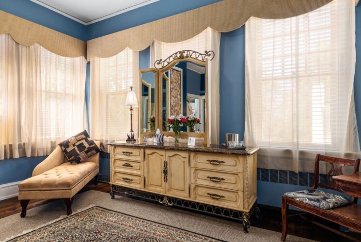 Bedroom with blue walls, hardwood flooring, light wooden antique dresser with mirror, tan upholstered chaise, and many large windows