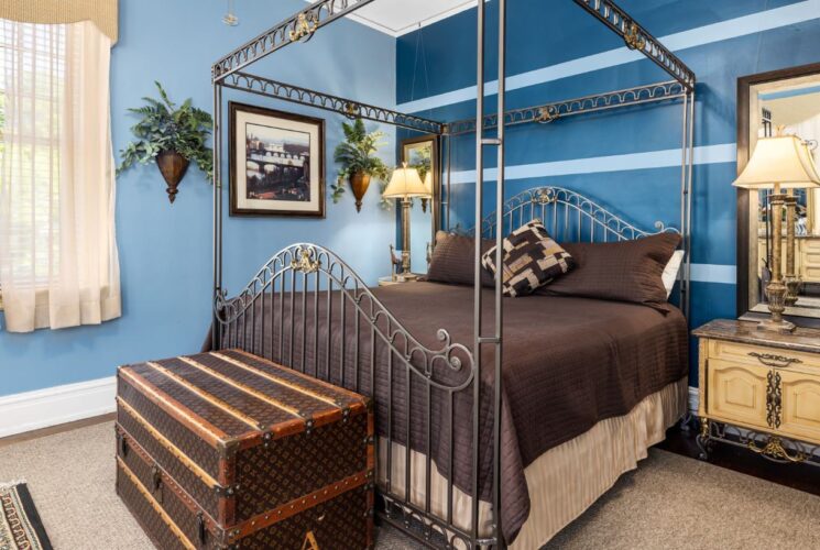 Bedroom with blue walls, hardwood flooring, wrought iron canopy bed, brown bedding, light wooden antique nightstand with lamp, and large brown trunk