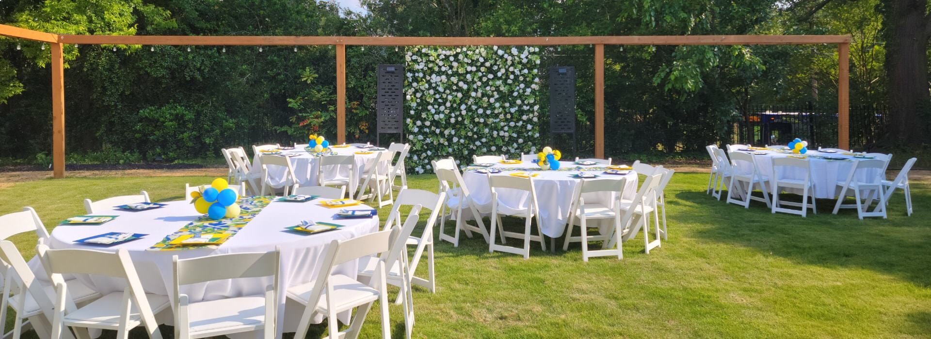 Backyard space set up for an event with multiple round tables with white tablecloths and white chairs set up on a green lawn surrounded by large trees