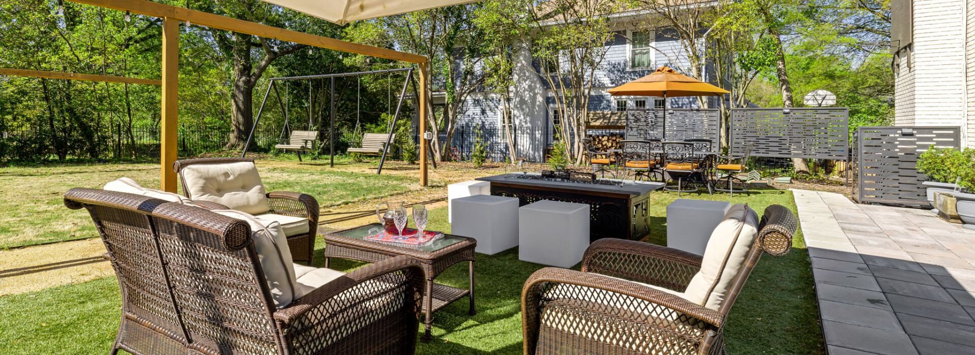 Backyard patio with brown wicker patio furniture, large tan umbrella, fire pit, and large green lawn