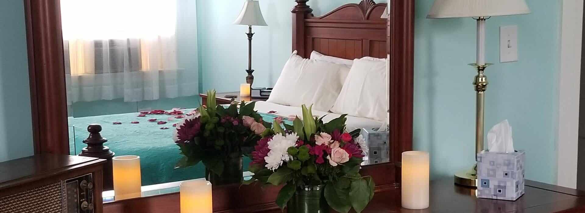 Close up view of large wooden mirror in bedroom with light blue walls reflecting a dark wooden bed with teal bedding and sprinkled with red and pink rose petals