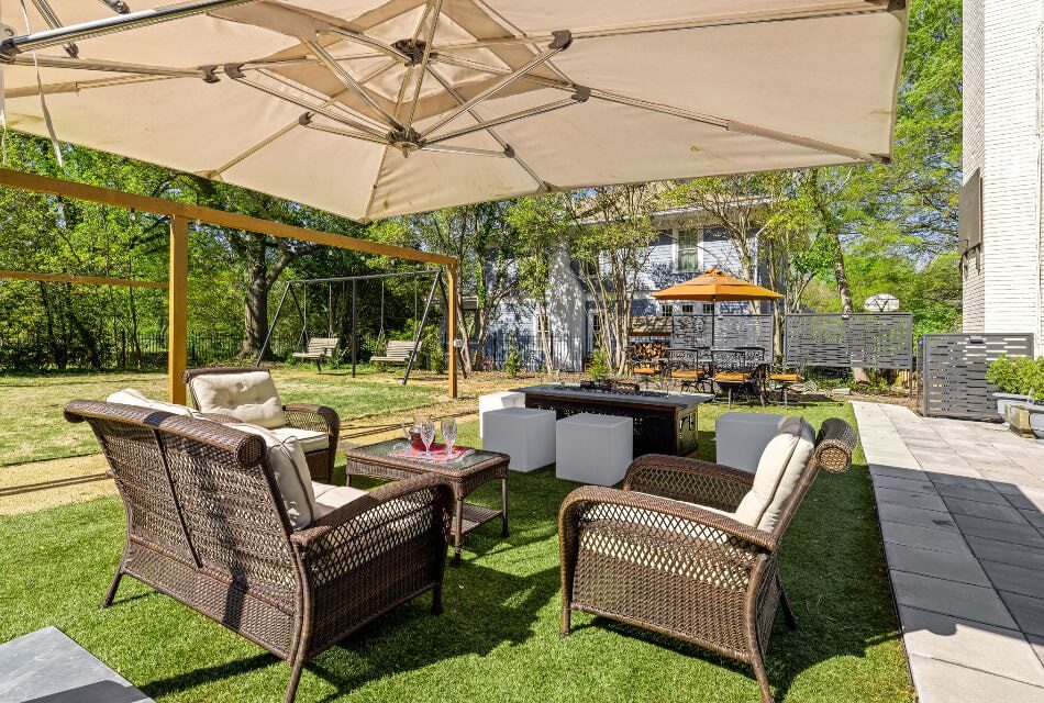 Backyard patio with brown wicker patio furniture, large tan umbrella, fire pit, and large green lawn