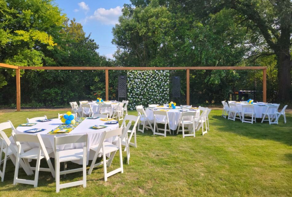 Backyard space set up for an event with multiple round tables with white tablecloths and white chairs set up on a green lawn surrounded by large trees