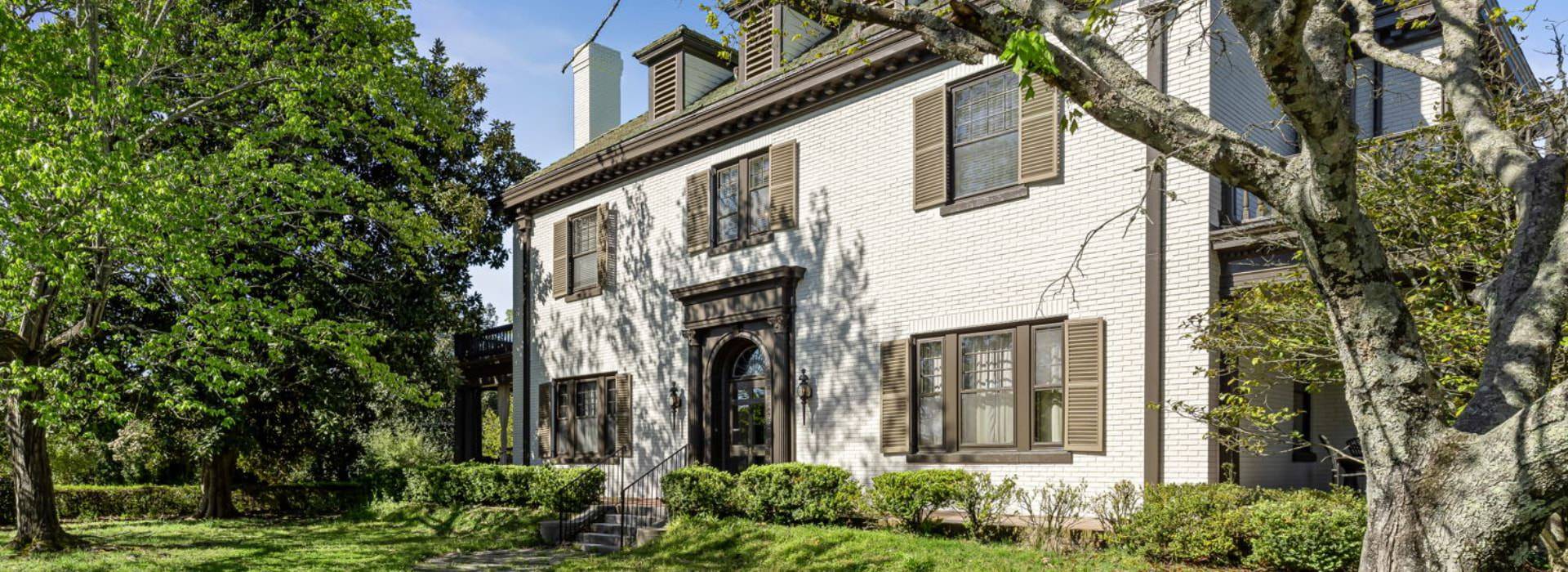 Exterior view of property painted white with brown trim and brown shutters and surrounded by green bushes, green lawn, and green trees