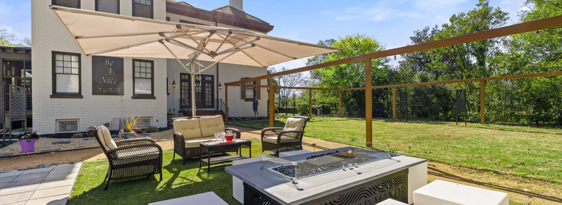 Backyard patio with brown wicker patio furniture, large tan umbrella, fire pit, and large green lawn