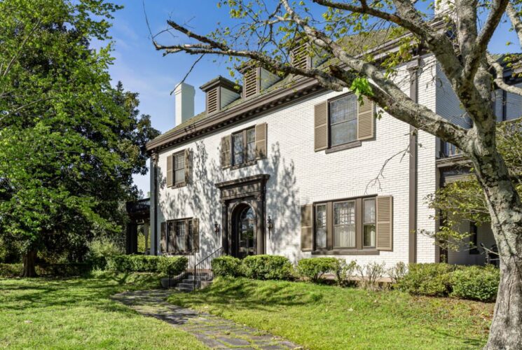 Exterior view of property painted white with brown trim and brown shutters and surrounded by green bushes, green lawn, and green trees