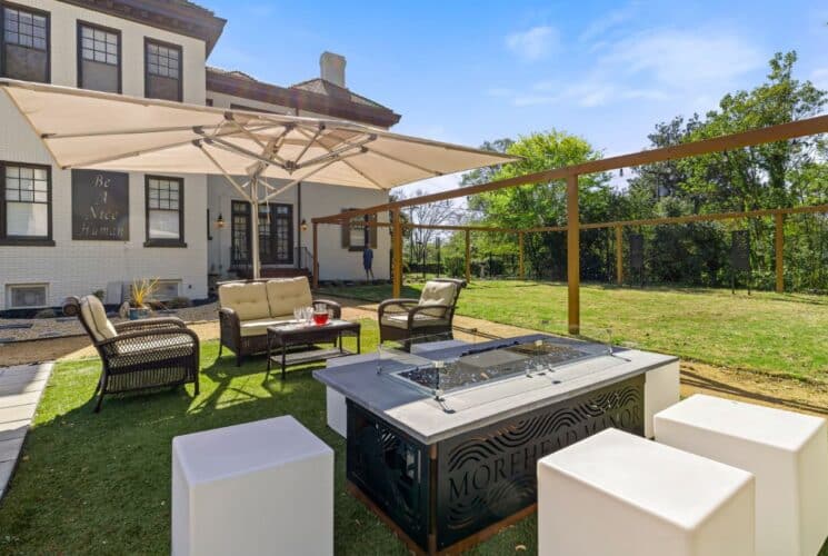 Backyard patio with brown wicker patio furniture, large tan umbrella, fire pit, and large green lawn